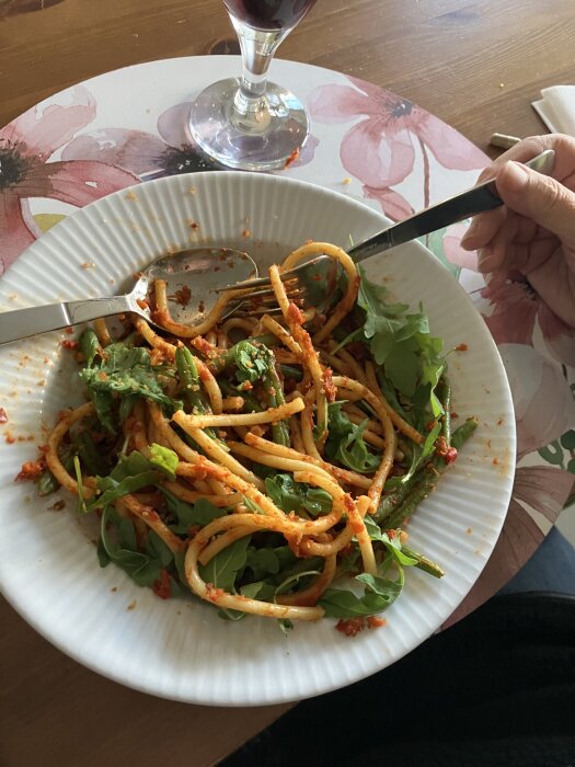 Tallrik med bucatini, haricot verts, ajvar, rucola och panko, serverad pÄ ett blommigt bordstablett med ett glas röd dryck i bakgrunden.