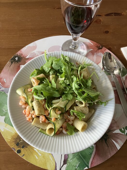 Tallrik med rigatoni, zucchini, rucola och krÀftstjÀrtar. Glas med röd dryck i bakgrunden.