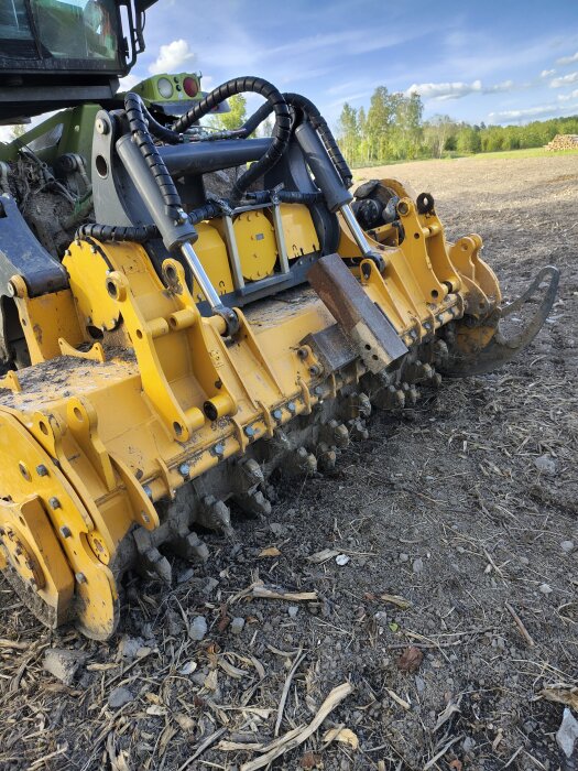 Maskin med stor fräs till stubbrytning på kalhygge, omgiven av stubbar och gräs, under en klar himmel.