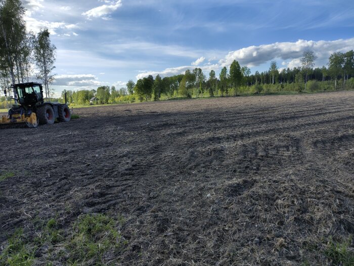 Ett öppet fält med trädridå i bakgrunden, en maskin till vänster på åkern under en klarblå himmel.