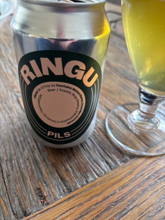 En burk öl märkt "RINGU Pils" och ett glas med öl på ett träbord.
