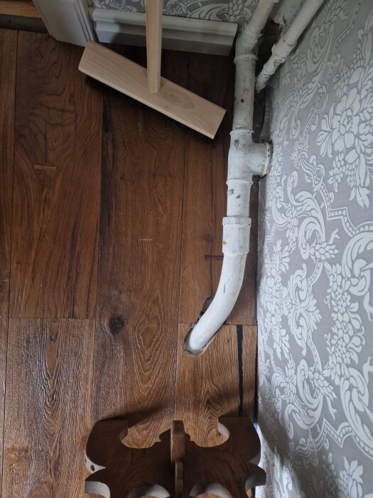 Gammalt rörsystem längs väggen med mörkt trägolv och mönstrad tapet i bakgrunden. En träborste står lutad mot väggen.