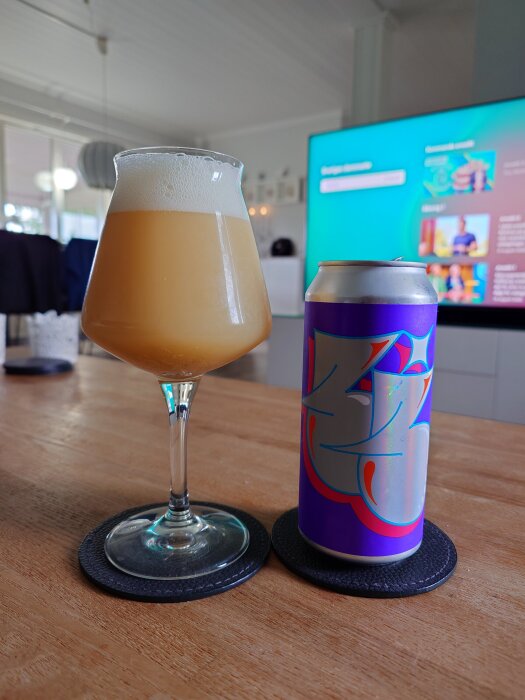 Glas med skummande öl och en färgglad ölburk från Omnipollo på ett bord framför en TV.