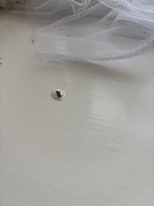 Pieni reikä fönsternischissa, jossa valkoista maalia ympärillä ja rei'itettyä kangasta yläpuolella. Mahdollista kosteusvauriota.