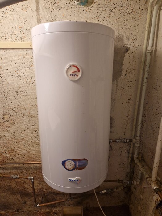 Varmvattenberedare från TESY monterad på en vägg med rör kopplade runtomkring i ett källarliknande utrymme.