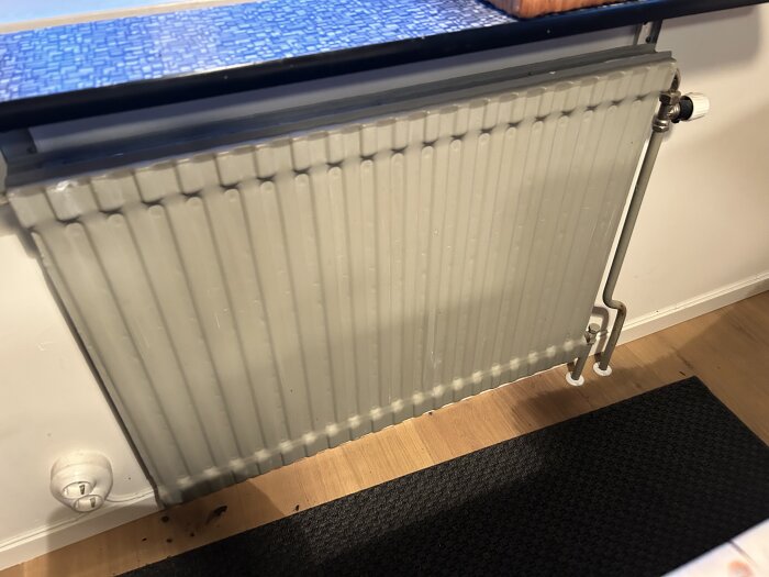 Grått väggmonterat element med synliga rör och termostat. Blått bord ovanför och mörk matta på trägolv.