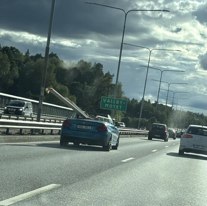 Blå bil på motorväg bärande en lång träbalk, nära Vallby-motet, under molnig himmel.