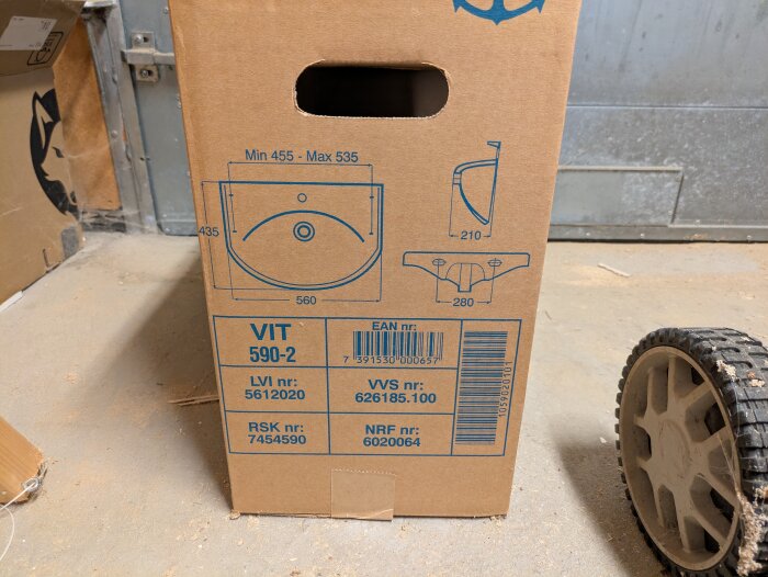 Kartong med tvättställ Gustavsberg Nordic 590-2, visar mått och produktinformation, står på garagets cementgolv bredvid ett hjul.