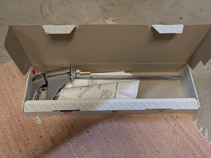 Kartong med en oanvänd Oras Safira Classica 282 blandare och tillhörande installationsmanualer, placerad på rosa mattan i ett garage.