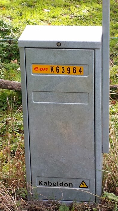 Grått elskåp med märket "e.on K63964" och texten "Kabeldon" längst ner, placerat i grönska.