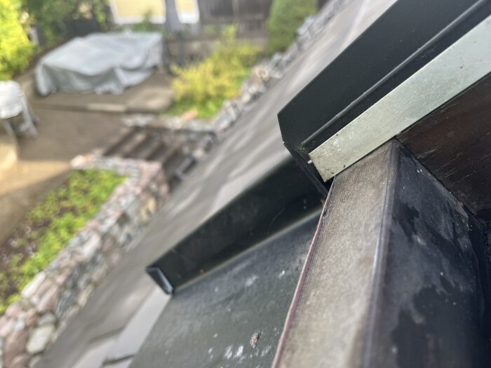 Närbild på en del av taket med en metallkant, i bakgrunden syns en trädgård med täckt objekt och terrass.