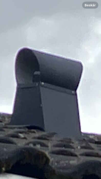 Takhuv i svart metall monterad på ett tegeltak, fotograferad utomhus under molnig himmel.