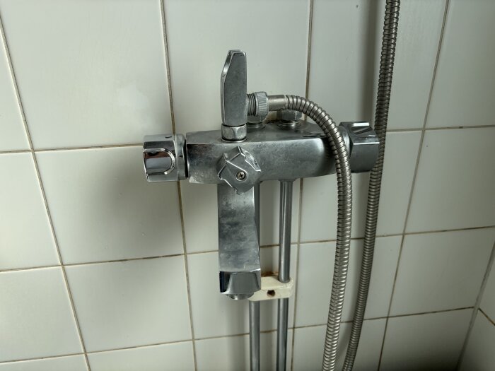 Duschblandare med trasigt reglage och koppar för dusch och pip visas framför kakelvägg.