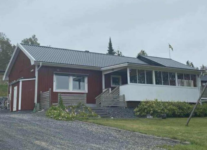 Rött trähus med grått tak, inglasad altan och grå trappa. Huset ligger i ett sommarstugeområde med en flagga synlig i bakgrunden.