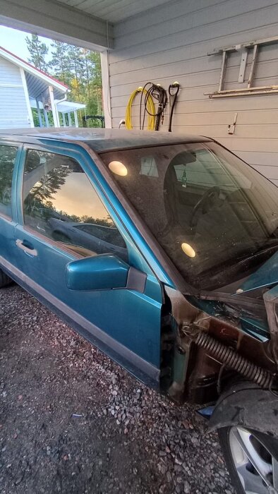 Blå bil med smutsiga vindrutetorkare och halvöppnad motorhuv i en carport, omgiven av verktyg och husvägg.