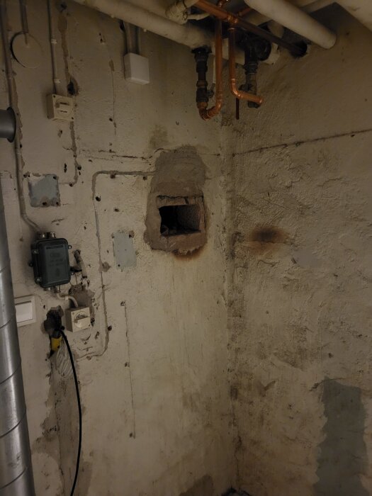 Hål i källarvägg efter borttagning av vattenburet värmesystem, med kopparrör och elkablar synliga på en grovputsad vägg.