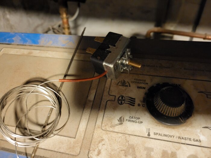 Trasigt vred och termometerkabel vid en gammal panna, med knappar och justeringsknapp synliga på panelen.