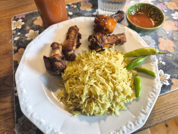 Tallrik med kycklingvingar, gult ris, gröna chilis och en skål med chilisås. Flaskor syns i bakgrunden.