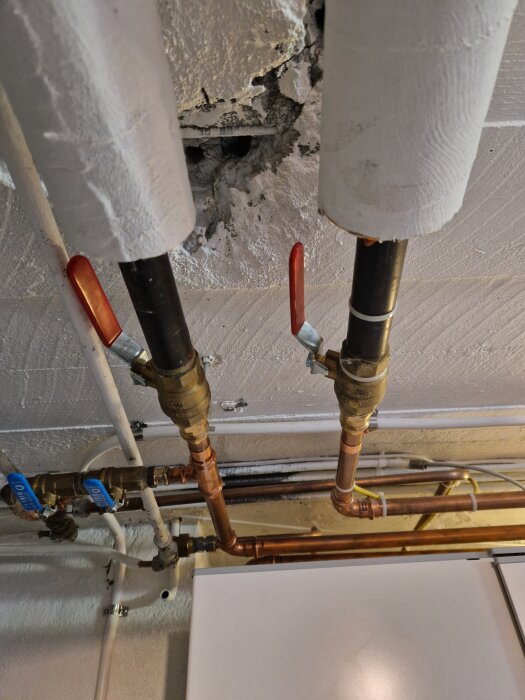 Vattenrör med ventiler och isolering i tak, synlig betongvägg med hål i bakgrunden.