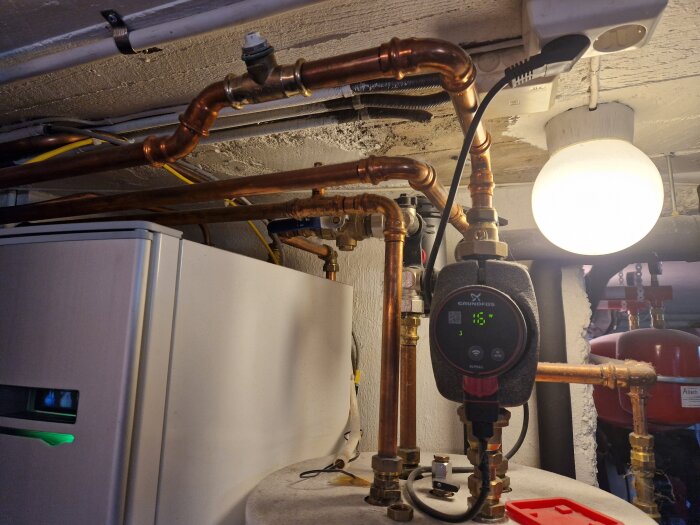 Värmepanna med kopparrör och cirkulationspump i en källare, belyst av en glödlampa.