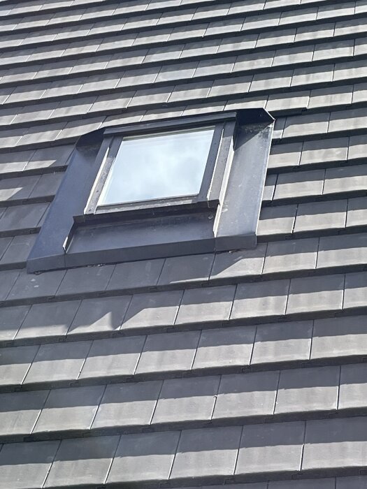 En bild av ett tak täckt med mörkgrå takpannor och ett fyrkantigt takfönster set utifrån.