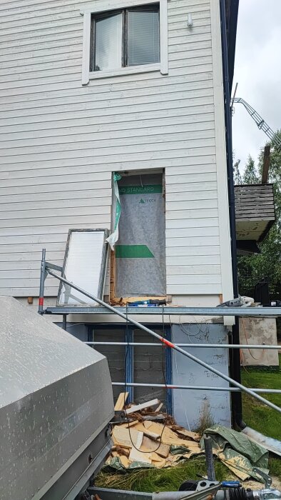 Öppning i husvägg för altandörr; brädfodring borttagen, byggställning och verktyg syns. Dörrkarm delvis monterad, byggmaterial nedanför.