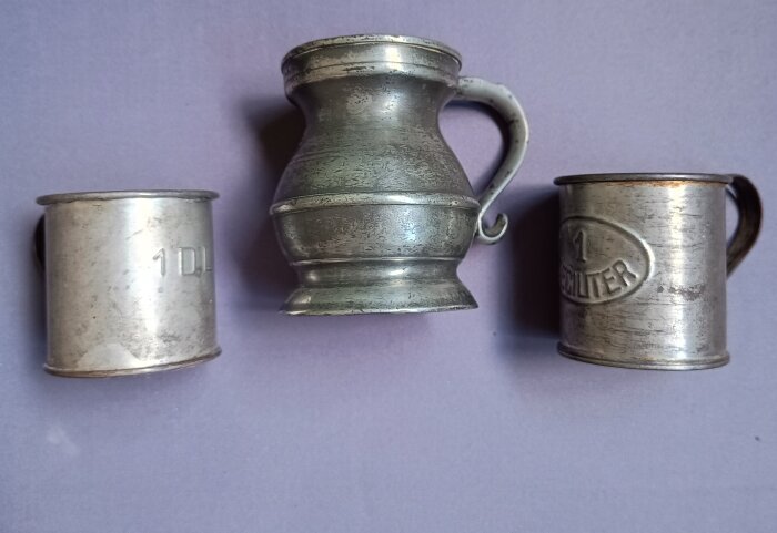 Tre äldre måttkärl i metall; till vänster och höger i bleckplåt och ett tennmått i mitten. Tennmåttet mäter en Gill (1/4 pint).