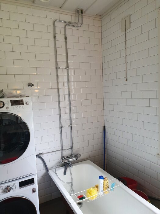 Badrum med vitt kakel, ett badkar med blandare och duschslang, och tvättmaskin ovanpå torktumlare. Takrörsynliga, ingen duschstång.
