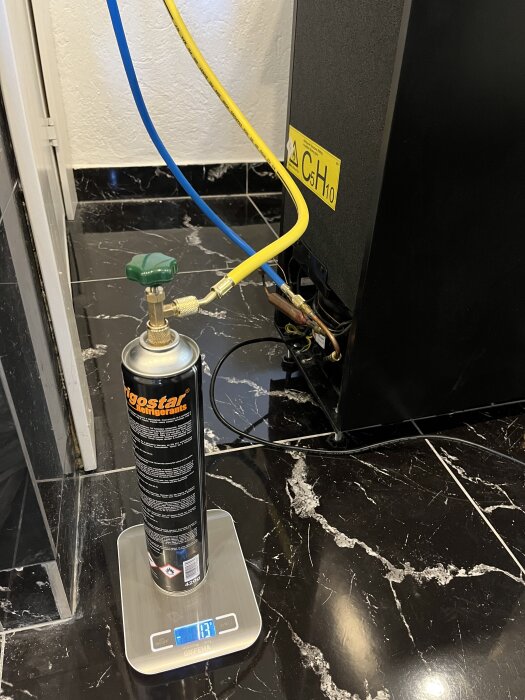 Gasflaska med grön ventil ansluten till blå och gul slang, placerad på våg, bredvid apparat med etikett "CSH10".