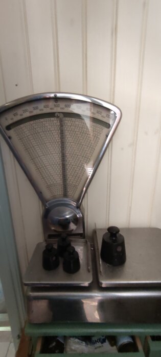 Gammaldags våg med vikter, placerad mot en panelvägg i ett kök eller förråd.