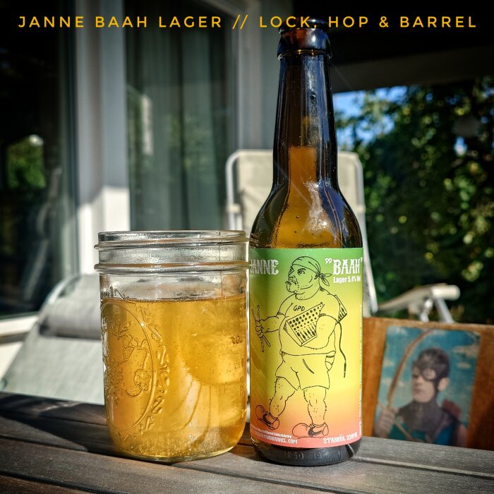 Lagerflaska och glasburk med öl på ett bord utomhus, etikett med tecknad figur och texten "Janne Baah Lager".