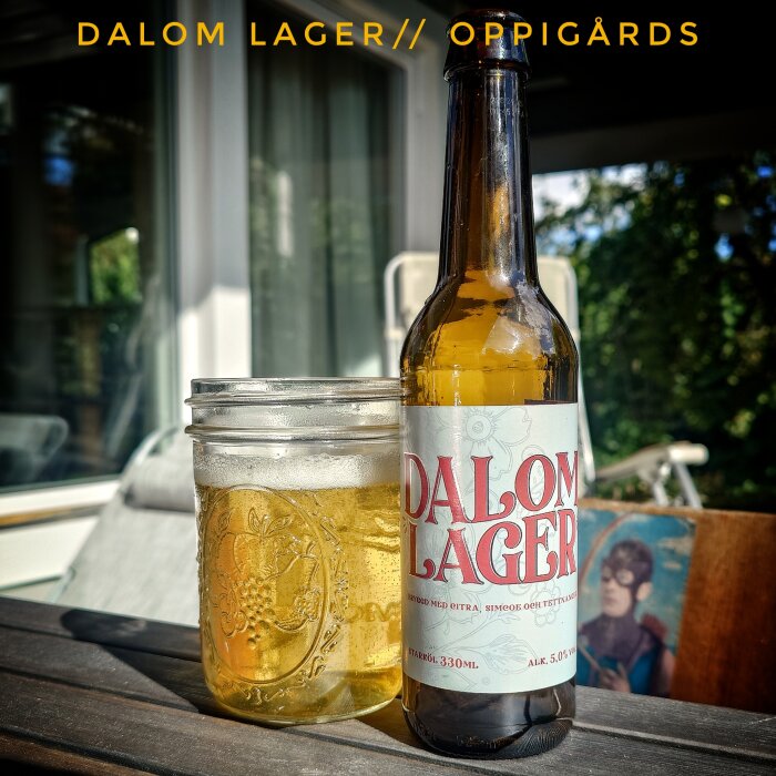 Flaska och glas med öl på utomhusbord, etikett med text "Dalom Lager", solbelyst miljö.