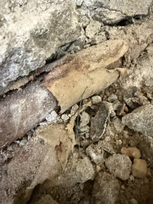 Ett gammalt rör med sliten isolering syns bland brutet källargolv. Isoleringen kan innehålla material som asbest.
