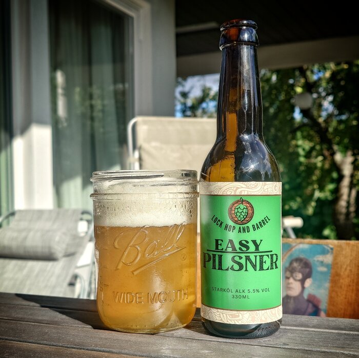 En flaska Easy Pilsner-öl och ett glas med öl står på ett bord utomhus framför en solstol.
