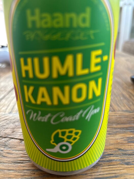 En närbild av en ölburk med etiketten "Haand Humle-kanon West Coast IPA" i gult på grön bakgrund, stående på ett träbord.
