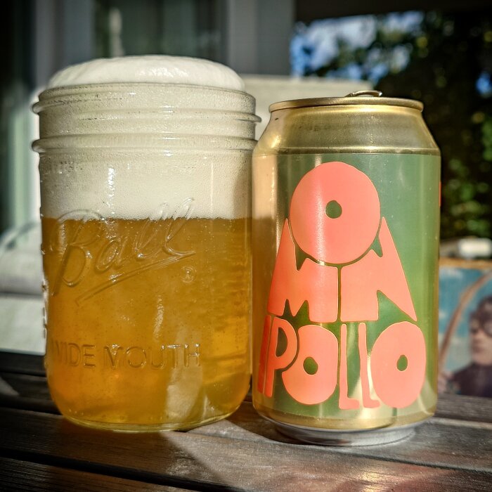 En burk Omnipollo pilsner och ett glas fyllt med öl med skumkrona, stående på ett bord utomhus.