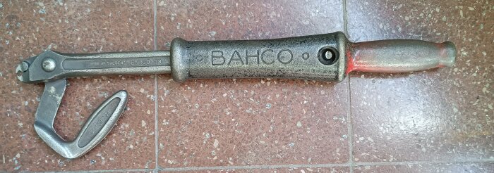Äldre modell Bahco 38, ett metallverktyg med texten "BAHCO" på handtaget, liggandes på ett rött golv med vita fläckar.