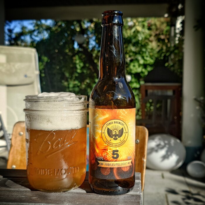 Ölflaska från Fenix Brewery och ett glas öl på ett bord utomhus, etikett med text "Jubileumslager" synlig.