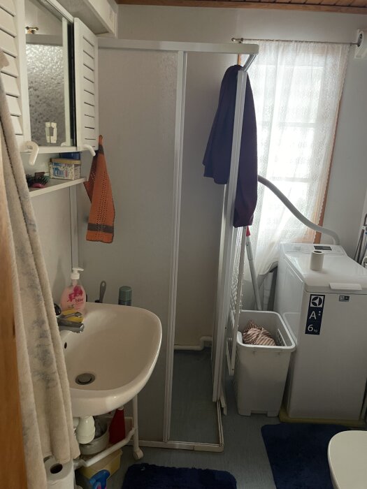 Badrum med handfat, spegel, orange handduk, duschkabin och tvättmaskin. En tvättkorg står bredvid ett fönster med spetsgardin.