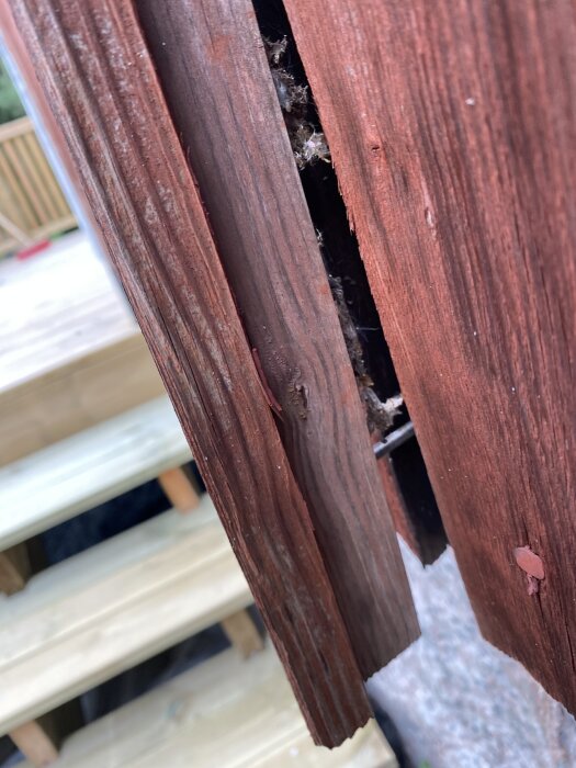 Rödbrun fasadbräda med lockläkt som har en träkil på undersidan, del av en gammal byggnad med trappsteg i bakgrunden.