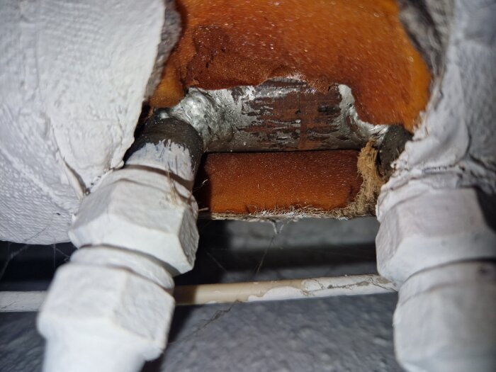 Närbild av rörkopplingar med orange isolering, omgivna av vitmålat material. Synliga rördetaljer och ytor.