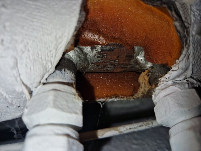 Närbild av två rör täckta med vit isolering och exponerade metallytor, med orangefärgat isoleringsmaterial synligt mellan rören.