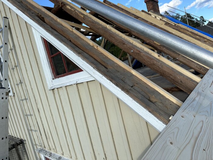 Takstolar förstärkta med nya reglar, synliga mellanrum mellan takstolar och yttervägg. Fönster på väggen under taklutningen, utsikt mot blå himmel.