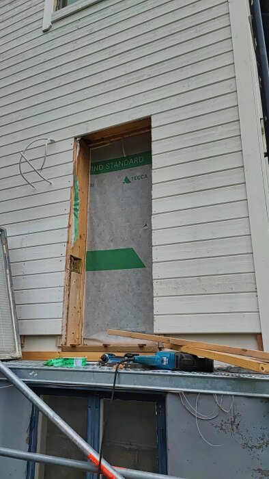 Öppning i husvägg för dörrkarm, omgiven av träreglar. Verktyg och material finns på byggställning nedanför.