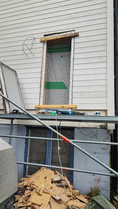 Byggarbetsplats med ett fönster utan glas, omgiven av verktyg och byggmaterial, samt en hög med avfall framför väggen.