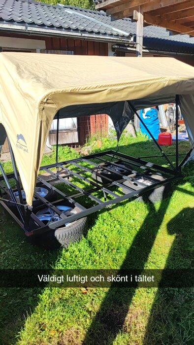 Tält med metallram står uppställt utomhus på en gräsmatta. Två hjul syns under ramen och ett tält i beige färg är monterat ovanpå.