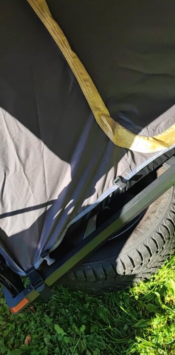 Tält fastsatt med hakar på en bilram, visar ett spänt tyg över ett däck, fotograferat i dagsljus på gräs.