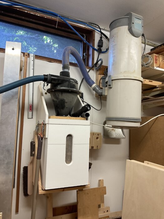 Centraldammsugaranläggning med cyklon, slangar och anslutna rör i ett hobbyrum. Cyklonen står ovanpå en vit låda bredvid centraldammsugaren.
