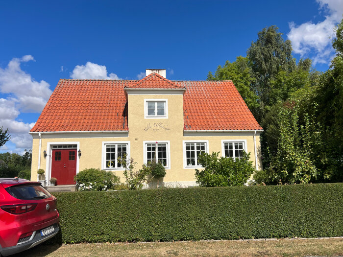 Gult hus med rött tegeltak och röda dörrar, två fönstren i mitten har spröjs och tolv rutor vardera.