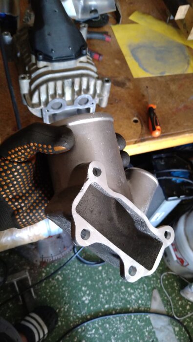 En hand i arbetshandske håller ett nygjutet aluminiumstycke framför en arbetsbänk med verktyg.